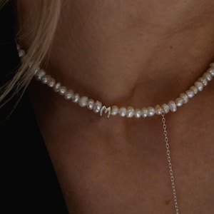 May include: A delicate pearl necklace with a silver charm in the shape of the letter "M". The necklace is on a model's neck and has a silver chain extending from the clasp.