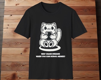 Op de afbeelding: Zwart T-shirt met een witte cartoonkat die een smartphone vasthoudt en de tekst "Waarom dromen najagen als je memes kunt scrollen?"