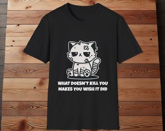 Op de afbeelding: Zwart T-shirt met een cartoonillustratie van een verdrietige, gebandageerde kat. De tekst "What doesn't kill you makes you wish it did" staat onder de kat.