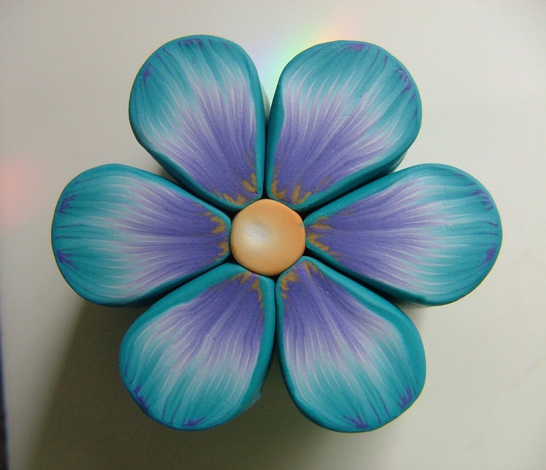 May include: A six-petal flower made of polymer clay. The petals are a gradient of teal and purple. The center of the flower is a light brown circle.