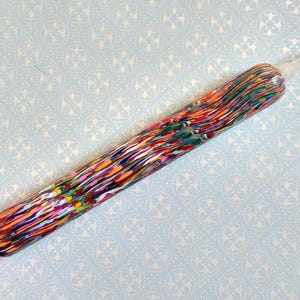 May include: A colorful pen with a swirling pattern of red, orange, yellow, green, and blue. The pen has a gold-colored tip and a clear plastic section near the tip. The pen is lying on a light blue surface.