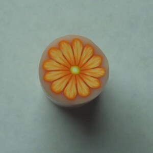 May include: A small, round, white polymer clay bead with an orange flower design. The flower has a yellow center and orange petals.