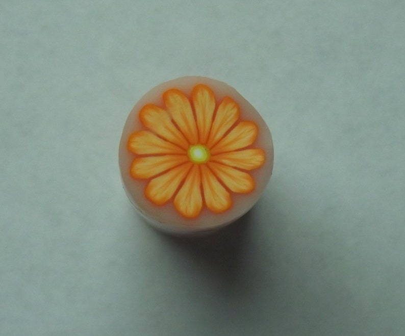 May include: A small, round, white polymer clay bead with an orange flower design. The flower has a yellow center and orange petals.