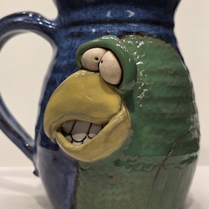 May include: A handmade ceramic mug with a parrot design. The mug features a blue exterior with a green parrot face, a yellow beak, and white teeth. The mug has a handle and is suitable for hot or cold beverages.