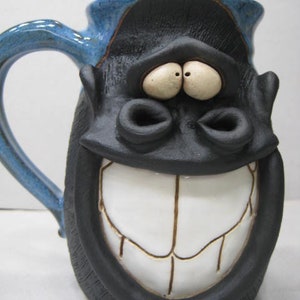 May include: A blue and black ceramic mug with a whimsical face. The mug has a wide, toothy grin and large, round eyes.
