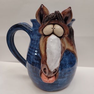 May include: A blue ceramic mug with a brown and white horse head sculpted on the front. The horse has a large, pink tongue sticking out.