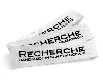 Könnte beinhalten: Drei weiße Stoffetiketten mit dem Wort "RECHERCHE" in Schwarz gedruckt. Der Text "HANDMADE IN SAN FRANCISCO" ist unter dem Wort "RECHERCHE" gedruckt. Die Etiketten sind gestapelt und abgewinkelt.