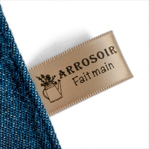 May include: A close-up of a beige fabric tag with the text "ARROSOIR Fait main" in black. A small illustration of a watering can with flowers is also on the tag. The tag is attached to a blue fabric.