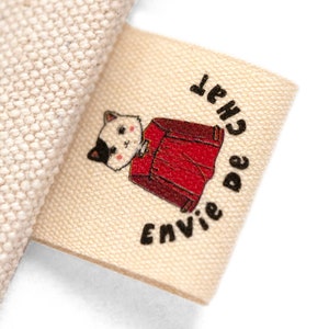 Può includere: Un'etichetta in tessuto beige con testo nero che recita "Envie de Chat" e un'illustrazione di un gatto dei cartoni animati che indossa una camicia rossa.