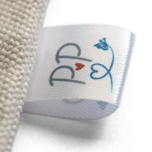 May include: Close-up of a white satin label with a logo. The logo features two stylized gray letter "P"s, a small red heart, and a blue floral design. The label is attached to a beige fabric.