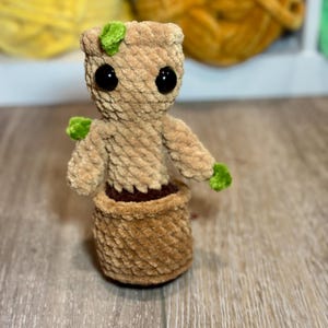 以下が含まれることがあります： 緑色の頭と大きな黒い目をした、植物のような小さな茶色の編みぐるみぬいぐるみ。茶色の編みぐるみの鉢に入っています。