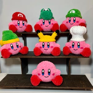 May include: A collection of handmade pink crochet plush toys, each with a smiling face and red feet. Some are wearing hats resembling characters like Mario, Pikachu, and a chef's hat. The toys are displayed on a dark wooden shelf.