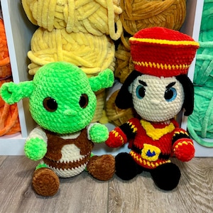 May include: Two handmade crochet dolls. One is green with large eyes and ears, wearing a brown vest. The other doll is dressed in red and black with a yellow collar and a red hat. Yarn in various colors is in the background.