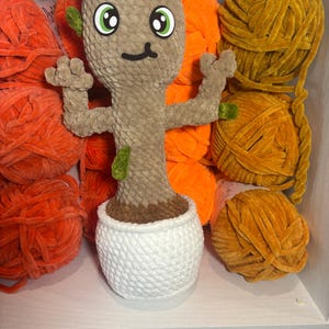 May include: A plush, light brown character with green accents, large eyes, and a smiling face, in a white pot. The character is surrounded by orange and yellow yarn balls, creating a whimsical display.