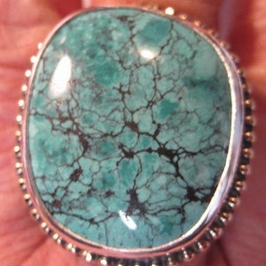 May include: A turquoise ring with a silver-colored bezel and a beaded edge. The turquoise stone has a mottled pattern of teal and black. The ring is a square shape with rounded corners.