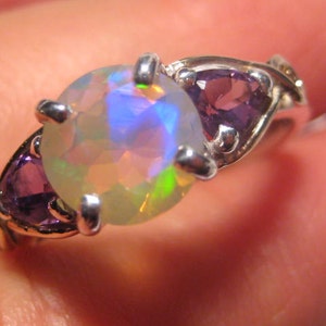 May include: A silver ring featuring a large, iridescent opal gemstone in the center, flanked by two smaller, triangular purple amethyst stones. The opal displays a play of colors, including blue, green, and yellow. The ring is set against a blurred background.
