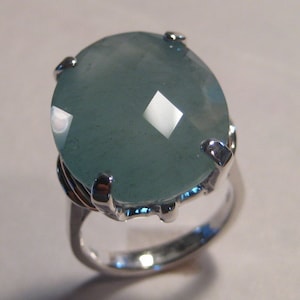 May include: A silver ring featuring a large, oval-cut, pale blue gemstone. The gemstone is faceted, catching the light and creating a sparkling effect. The ring's setting has a claw-like design, securely holding the stone.