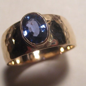 May include: A gold ring with a hammered texture and a large, oval-cut blue gemstone. The gemstone is set in a bezel setting, and the band is wide and polished. The ring is shown at an angle, highlighting the details of the design.