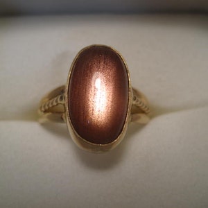 May include: A gold ring featuring a large, oval-shaped, reddish-brown gemstone. The ring's band has a textured design on the sides, and the gemstone is set in a gold bezel. The ring is displayed on a white surface.