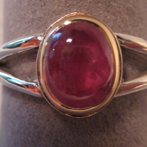 May include: An oval-shaped ruby gemstone ring with a gold bezel setting and a split silver band. The ruby has a deep red color and is the focal point of the ring. The ring is displayed on a gray surface.