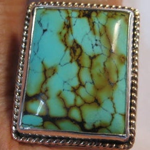 May include: A square turquoise ring with a silver-colored rope-like border. The turquoise stone has a light blue-green hue with brown veining. The ring is a statement piece, showcasing the natural beauty of the stone.