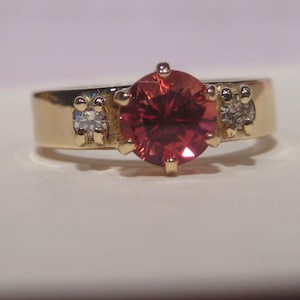May include: A gold ring featuring a large, round, red gemstone as the centerpiece. Two smaller, clear gemstones are set on either side of the main stone. The ring has a classic, elegant design.