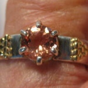 May include: A close-up of a ring featuring a peach-colored gemstone set in a silver and gold band. The gemstone is held in place by six silver prongs. The band has gold decorative elements on each side.