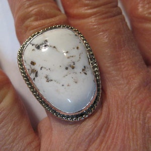 May include: A large, oval-shaped ring featuring a white stone with brown and black speckles. The stone is set in a silver-colored metal band with a braided rope design around the edge. The ring is on a finger.
