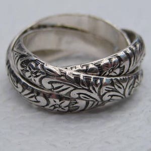 May include: A silver ring with an intricate floral design. The ring is composed of three interlocking bands, each adorned with detailed carvings of flowers and leaves. The ring has a polished, reflective surface.