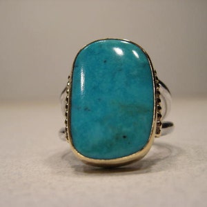 May include: A silver ring with a large, oval turquoise stone set in a gold bezel. The ring has a double band and decorative detailing on the sides of the stone. The turquoise is a vibrant blue-green color.