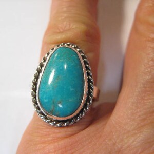 May include: A silver ring featuring a large, oval turquoise stone. The stone is a vibrant blue-green color and is set in a silver bezel with a twisted rope design around the edge. The ring is on a finger.