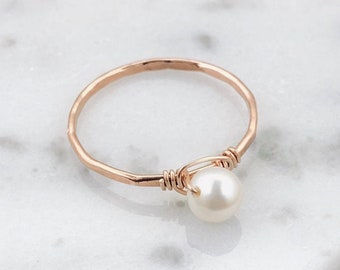 May include: A delicate rose gold ring featuring a single, lustrous white pearl. The ring's band has a hammered texture, and the pearl is secured with wire wrapping. The ring is displayed on a white marble surface.