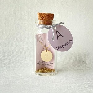 Puede incluir: Un collar de oro relleno con un colgante circular grabado con la letra "A" se muestra dentro de una pequeña botella de vidrio. La botella está sellada con un corcho y una etiqueta que dice "A" y "14k gold-fill".