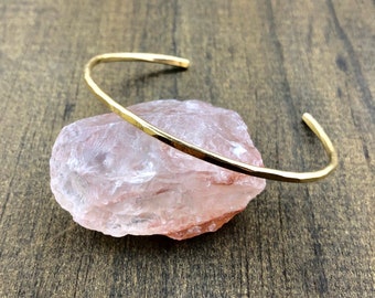 May include: A gold-colored, hammered-finish cuff bracelet rests on a light pink, rough-textured stone. The bracelet has a simple, open design. The background is a dark brown, wood-grain surface.
