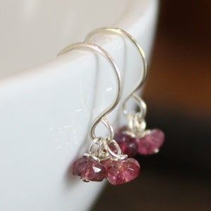 May include: A pair of dangle earrings with silver-colored hooks. Each earring features three small, faceted, pink gemstones. The earrings are displayed against a white background, with a blurred brown background.