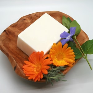 May include: A rectangular white bar of soap resting in a natural, irregular-shaped wooden bowl. The soap is accompanied by two bright orange calendula flowers, a single purple periwinkle flower, and small green leaves tucked into the bowl.