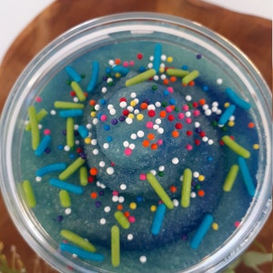 May include: A clear circular container filled with a blue, granular substance swirled into a peak. The surface is decorated with small, multi-colored round sprinkles and larger, cylindrical sprinkles in shades of lime green and bright blue.