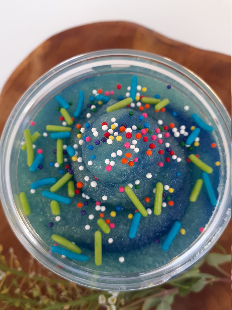 May include: A clear circular container filled with a blue, granular substance swirled into a peak. The surface is decorated with small, multi-colored round sprinkles and larger, cylindrical sprinkles in shades of lime green and bright blue.