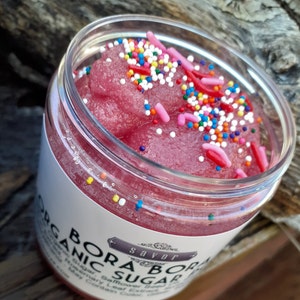 May include: A clear plastic jar filled with a pink, granular sugar scrub topped with colorful round sprinkles and pink rod-shaped sprinkles. The white label on the jar features the text BORA BORA ORGANIC SUGAR and the brand name savor in a decorative frame.
