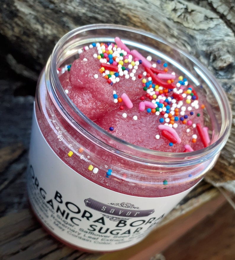 May include: A clear plastic jar filled with a pink, granular sugar scrub topped with colorful round sprinkles and pink rod-shaped sprinkles. The white label on the jar features the text BORA BORA ORGANIC SUGAR and the brand name savor in a decorative frame.