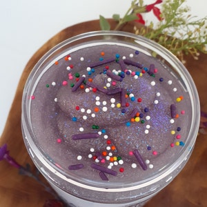 May include: A clear plastic jar filled with a thick, purple, glittery substance resembling slime or scrub. The surface is topped with small, round, multi-colored sprinkles and larger, cylindrical, dark purple sprinkles scattered throughout.