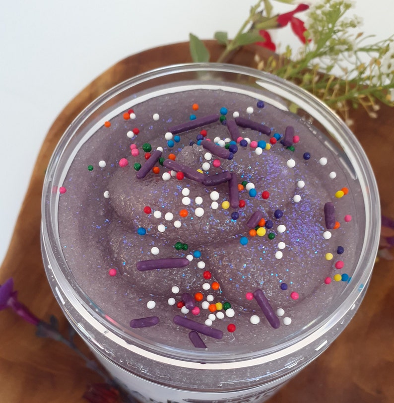 May include: A clear plastic jar filled with a thick, purple, glittery substance resembling slime or scrub. The surface is topped with small, round, multi-colored sprinkles and larger, cylindrical, dark purple sprinkles scattered throughout.