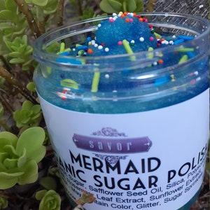 May include: A clear plastic jar filled with blue granulated sugar polish topped with colorful sprinkles. The white label features the text Savor Mermaid Sugar Polish and lists ingredients including organic sugar, safflower seed oil, and silica.