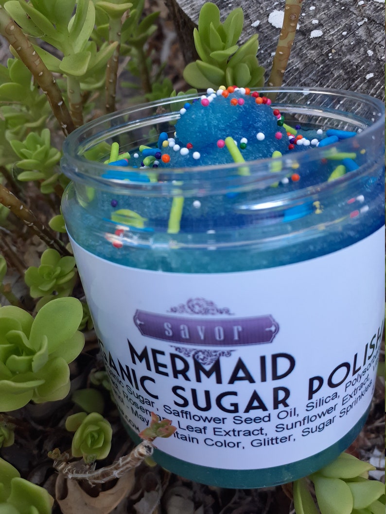 May include: A clear plastic jar filled with blue granulated sugar polish topped with colorful sprinkles. The white label features the text Savor Mermaid Sugar Polish and lists ingredients including organic sugar, safflower seed oil, and silica.
