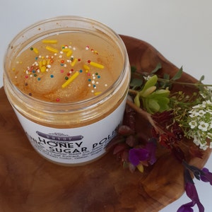 May include: A clear plastic jar filled with golden honey sugar polish, topped with colorful sprinkles. The white label features the text Savor Honey Sugar Polish. The jar is placed on a wooden surface next to small white and purple flowers.