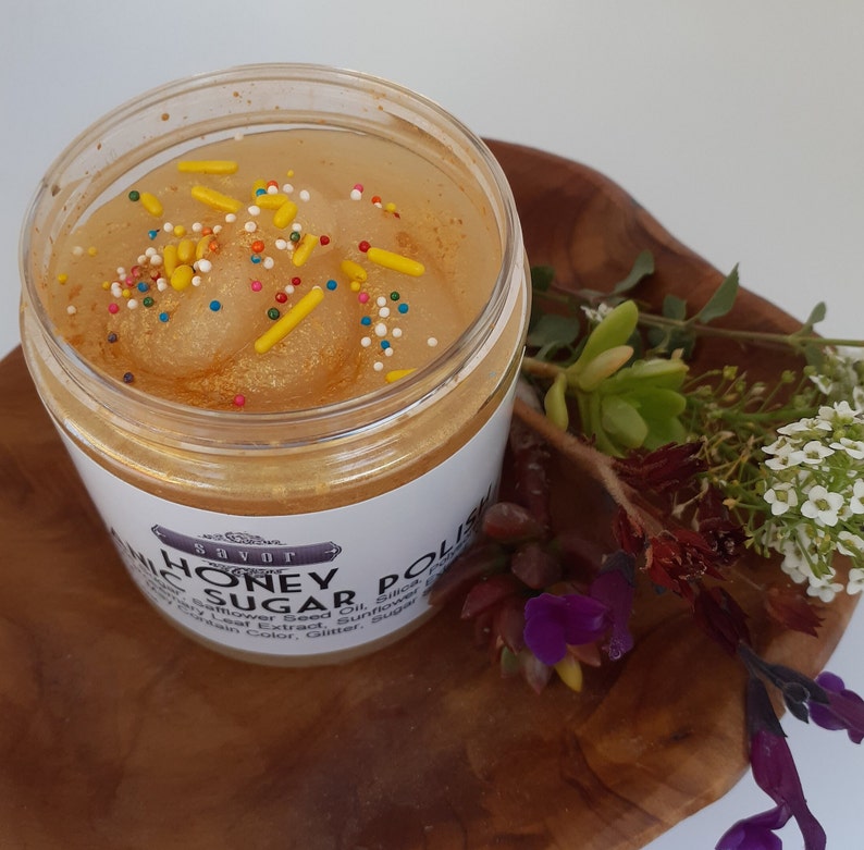May include: A clear plastic jar filled with golden honey sugar polish, topped with colorful sprinkles. The white label features the text Savor Honey Sugar Polish. The jar is placed on a wooden surface next to small white and purple flowers.