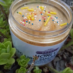 May include: A clear plastic jar filled with a golden-toned sugar scrub, topped with yellow rod-shaped sprinkles and small multicolored round sprinkles. The white label on the jar features the text Savor Organic Honey Sugar in black font.