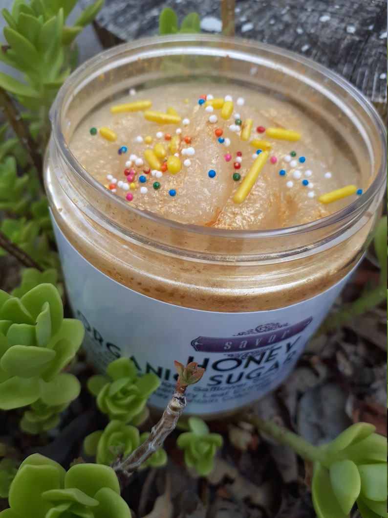 May include: A clear plastic jar filled with a golden-toned sugar scrub, topped with yellow rod-shaped sprinkles and small multicolored round sprinkles. The white label on the jar features the text Savor Organic Honey Sugar in black font.