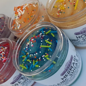 May include: Five clear plastic jars filled with colorful sugar scrubs topped with multi-colored sprinkles. The jars feature white labels with the brand name Savor and product names including Honey Organic Sugar and Mermaid Polish Organic Sugar.