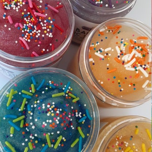 May include: Four clear plastic jars filled with colorful, textured slime. Each jar features a distinct color: deep pink, dark purple, bright orange, and teal blue. The surface of each slime is topped with a variety of small, multi-colored sprinkles.